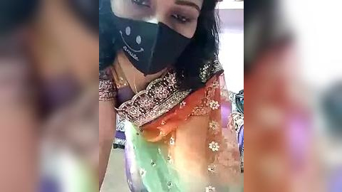 Media: A video of a South Asian woman wearing a colorful sari with intricate embroidery, a black face mask with a smiling emoji, and a blurred background.