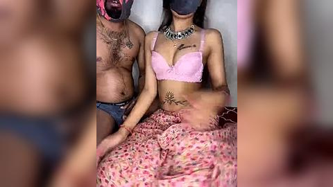 Media: Video of a man with a beard and tattoos wearing a black mask and blue shorts, and a woman with long black hair, pink bra, and floral skirt, sitting on a bed, both with face masks.