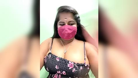 Media: Video of a South Asian woman with medium brown skin, long black hair, and a red face mask, wearing a black floral dress with low-cut neckline, posing against a green wall, blurry background.