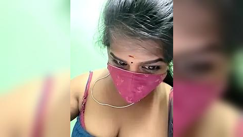 Media: Video of a South Asian woman with medium brown skin, dark hair, and a pink surgical mask. She wears a red spaghetti-strap top and has a noticeable red bindi on her forehead. The background is blurred, green and beige.