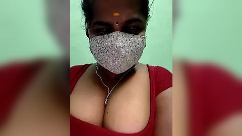 Media: Video of a South Asian woman with medium skin tone and dark hair, wearing a red top, a silver mask, and a bindi, set against a blurred background.