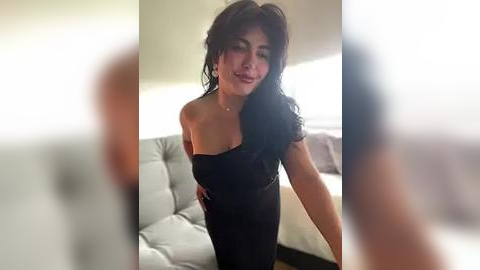 Media: Video of a smiling woman with long, wavy black hair, wearing a black off-shoulder dress, standing in a modern living room with white couches.