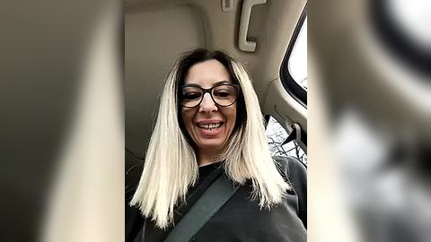 Media: Video of a woman with long, straight, platinum blonde hair and glasses, smiling in a car, wearing a black jacket, with a seatbelt across her chest, behind the steering wheel.