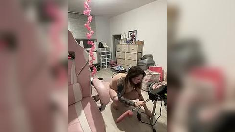 Media: Video of a young woman with tattoos, wearing a pink bra and panties, kneeling on a carpeted floor in a cluttered bedroom, surrounded by pink decorations and a video camera.