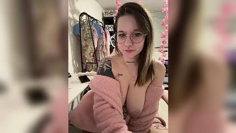 Media: Video of a young woman with glasses, light skin, and brown hair, wearing a pink fuzzy robe, showing cleavage, in a room with pink decorations, hanging clothes, and a mirror.