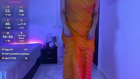 Media: A video of a person wearing a bright orange saree, standing in a dimly lit room with purple LED strip lighting. The person's face is not visible.