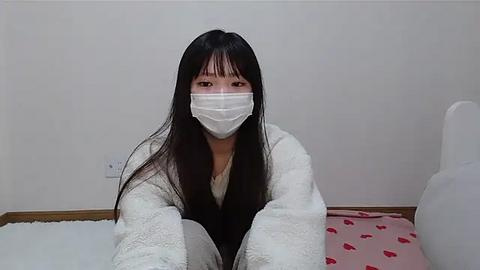 Media: Video of an Asian woman with long black hair, wearing a white face mask, a white bathrobe, and a pink bedspread with heart patterns. She sits on a white bed against a plain white wall, looking forward.