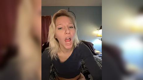 Media: Video of a blonde woman with a shocked expression, wearing a dark top, sitting on a bed with patterned bedding. Background includes a lamp, wall, and blurred figures.