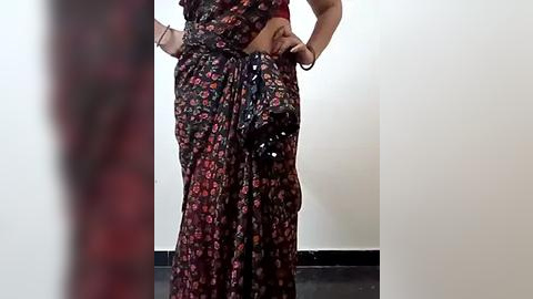 Media: Video of a person in a long, dark floral dress with a black floral pocket, standing with hands on hips, in a plain, white-walled room with black floor.