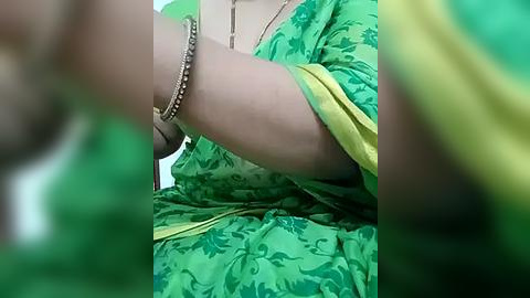 Media: Video of a woman wearing a green, floral-patterned sari with a yellow border, gold jewelry, and a bracelet, sitting with crossed legs. The background is blurred, emphasizing the sari's intricate design.