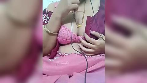 Media: A blurry video of a woman with fair skin, wearing a pink lace bra and matching skirt, with a gold necklace and bracelet, holding a phone, blurred background.