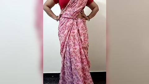 Media: Video of a woman in a vibrant pink and red floral sari with a red blouse, hands on hips, against a plain white wall and dark floor.