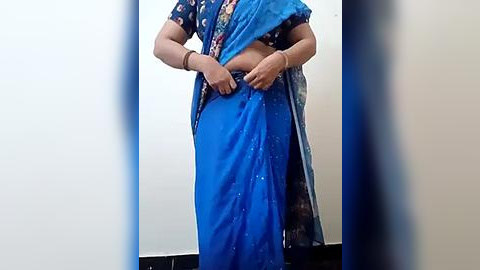 Media: A video of a woman in a blue sari, adjusting her sari with both hands, against a plain white wall.