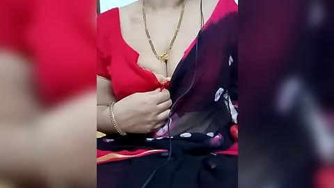 Media: Video of a woman's chest in a red blouse, partially unbuttoned, revealing cleavage. She wears a gold necklace and a black sari with floral patterns. The background is blurred.