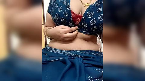 Media: Video of a woman in a blue saree with white polka dots, showcasing her cleavage in a red bra, wearing a gold necklace, against a blurred background.