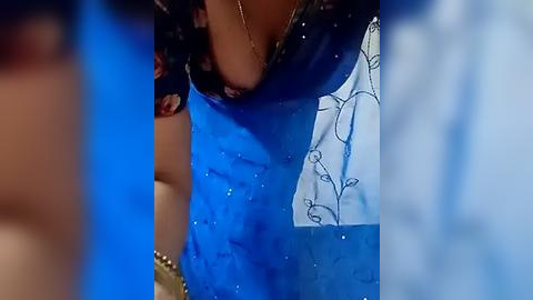 Media: A close-up video of a woman's upper body, wearing a blue saree with floral embroidery, and a black blouse with a low neckline, revealing her small breasts. The background is blurred, emphasizing the saree's vibrant colors and intricate details.