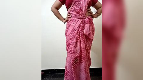 Media: Video of a person in a pink, pleated saree with a matching blouse, standing against a plain white wall. The background is blurred, focusing on the saree's texture and folds.