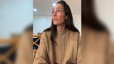 Media: Video of a woman with long dark hair in a beige knitted sweater, sitting indoors with blurred background.