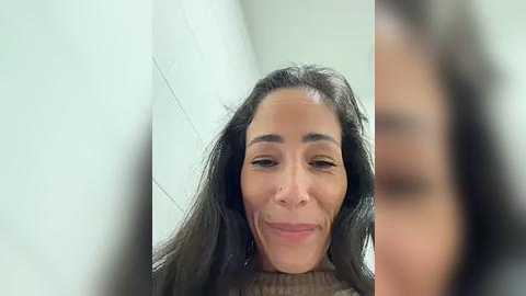 Media: Video of a woman with long, dark hair, light skin, and a wide smile, standing against a white tiled wall, slightly out of focus.