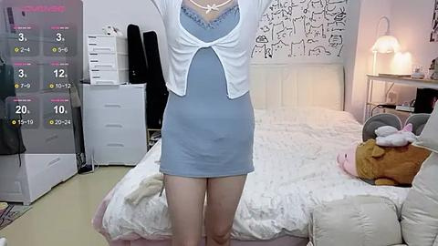 Media: A video of a young woman with fair skin, wearing a light blue mini dress, white cardigan, and a pink bra, standing in a cozy, white-themed bedroom with a bed, stuffed animals, and a lamp.