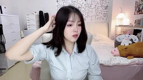 Media: Video of an East Asian woman with shoulder-length black hair, in a light blue shirt, adjusting her hair in a cozy bedroom with white walls, a bed, and stuffed animals.