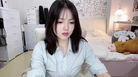 Media: Video of a young Asian woman with shoulder-length black hair, wearing a light blue button-down shirt, sitting on a white chair in a cozy, cluttered bedroom with a bed, stuffed animals, and white shelves.