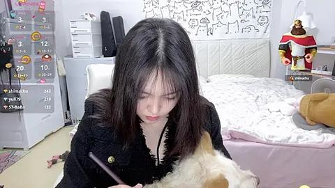 Media: Video of an Asian woman with long black hair, wearing a black dress, sitting on a bed, feeding a small dog. The room features white walls with a patterned tapestry, a plush toy, and a teddy bear.