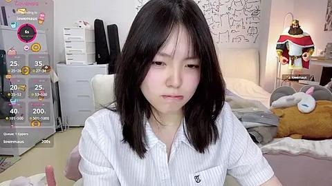 Media: Video of an Asian woman with shoulder-length black hair, wearing a white striped shirt, sitting in a cozy bedroom with a bed, stuffed animals, and a desk.