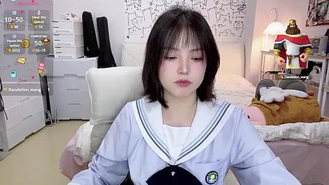 Media: Video of a young Asian woman with straight black hair, wearing a Japanese schoolgirl uniform, sitting in a cozy, white-walled bedroom with plush toys, a desk, and a lamp.