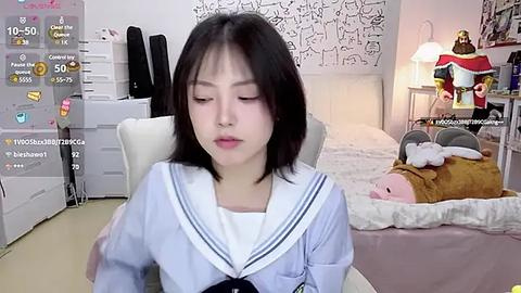 Media: Video of a young Asian woman with straight black hair, wearing a Japanese schoolgirl uniform, sitting on a white chair in a cozy bedroom with a teddy bear, white bedding, and a lamp.