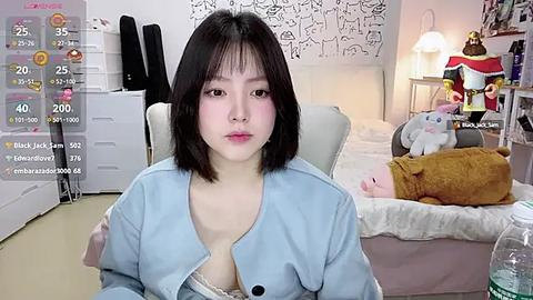 Media: A video of an Asian woman with shoulder-length black hair, wearing a light blue cardigan, sitting on a white chair in a cozy bedroom with white walls, a bed, and stuffed animals.