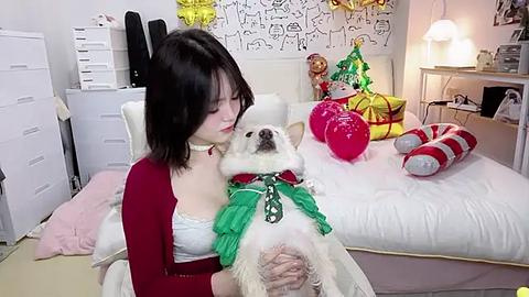 Media: Video of a young woman with shoulder-length black hair, wearing a red cardigan and white lace top, holding a white poodle dressed as Santa Claus in a festive bedroom with holiday decorations.