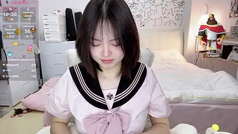 Media: A video of a young East Asian woman with straight black hair, wearing a pink sailor school uniform, sitting at a desk in a tidy, white bedroom with a plush toy and a lamp.