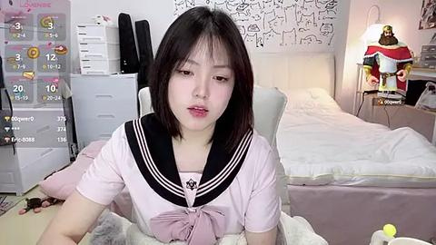 Media: Video of an East Asian woman with straight black hair, wearing a pink sailor school uniform, sitting on a bed with a white duvet and pillows, in a tidy, brightly-lit bedroom with a calendar, lamp, and decorative items.