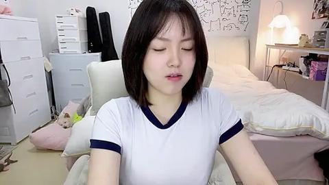 Media: Video of an Asian woman with straight black hair, wearing a white T-shirt with navy trim, sitting in a cluttered bedroom with white furniture, stuffed toys, and a bed.