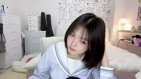 Media: Video of an Asian woman with straight black hair, wearing a sailor school uniform, sitting on a white bed in a cluttered, minimalist bedroom with white walls and a black-and-white abstract art piece.