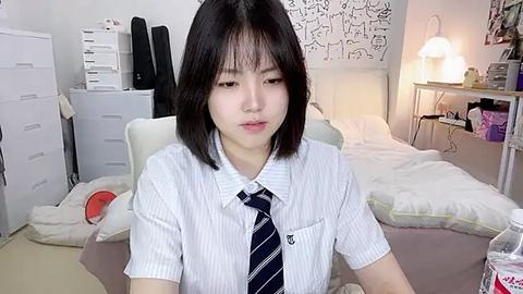 Media: Video of an East Asian woman with shoulder-length black hair, wearing a white striped shirt and blue tie, sitting at a desk in a minimalist, cluttered bedroom with white walls and a bed.