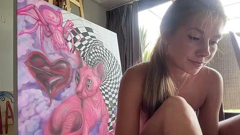 Media: Video of a topless young woman with fair skin and long brown hair, sitting in a room with a large, colorful, surreal painting featuring a pink cat and heart shapes.