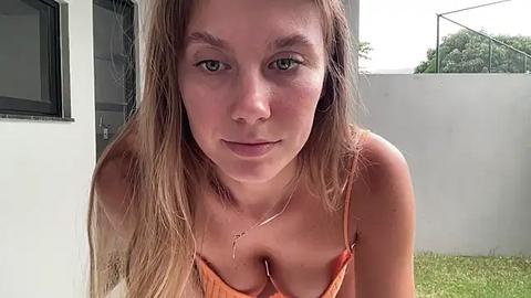 Media: Video of a young Caucasian woman with light skin and long, straight blonde hair, wearing an orange spaghetti-strap top. She has a neutral expression and is indoors near a window.