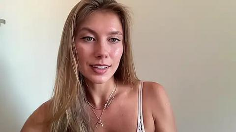 Media: Video of a young, light-skinned woman with long, straight blonde hair, wearing a white spaghetti-strap top, and silver necklace, against a plain, light beige wall.