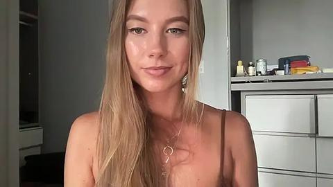 Media: Video of a topless Caucasian woman with long blonde hair, fair skin, and slender physique, wearing a necklace, standing in a modern, minimalist bathroom with grey cabinets and toiletries.