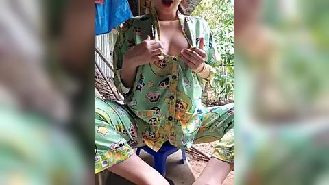 Media: A video captures a woman in a green pajama set with cartoon animals, seated outdoors, holding her shirt open to reveal cleavage. The background features blurred foliage and a blue tarp.