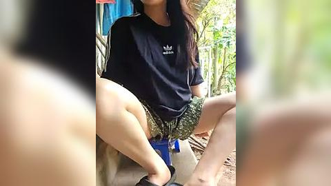 Media: Video of an Asian woman with long hair, wearing a black Adidas t-shirt and camouflage shorts, sitting on a blue bucket, outdoors, surrounded by greenery.