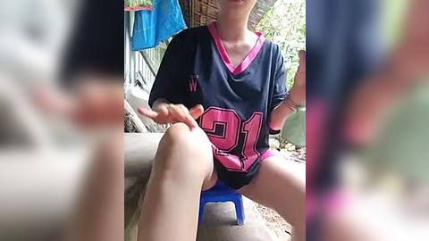 Media: Video of a young person in a navy-blue jersey with pink and white accents, sitting on a blue plastic chair, legs apart, outdoors near a wooden structure.