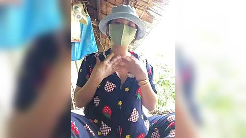 Media: Video of a woman in a blue dress with strawberry pattern, wearing a face mask, sitting outdoors under a thatched roof, holding her hands up.