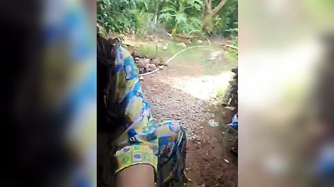 Media: Video of a person wearing a colorful, patterned shirt sitting on a dirt path in a dense, green forest, surrounded by tree trunks and foliage.