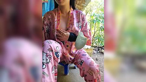 Media: Video of an Asian woman with medium build, light skin, and long dark hair, wearing a pink Hello Kitty pajama set, sitting outdoors, holding a gray cat toy, surrounded by green foliage and a blue tent.