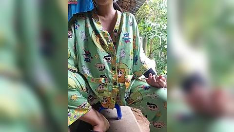 Media: A video of a person in a green, cartoon panda-patterned pajama set, seated on a rustic bench in a lush, tropical outdoor setting. The background features a thatched roof and dense green foliage.