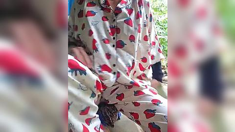 Media: A video of a person in a white pajama set with red strawberry print, sitting on the ground outdoors with blurred green foliage in the background.