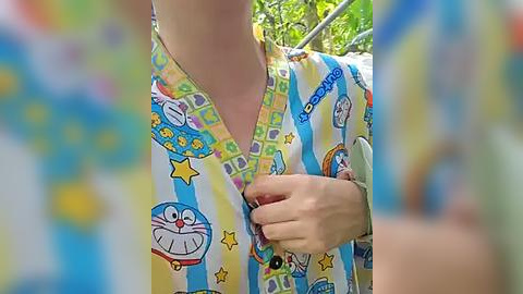 Media: Video of a person wearing a blue and yellow striped, cartoon-themed nightgown featuring Doraemon characters, with blurred background foliage.